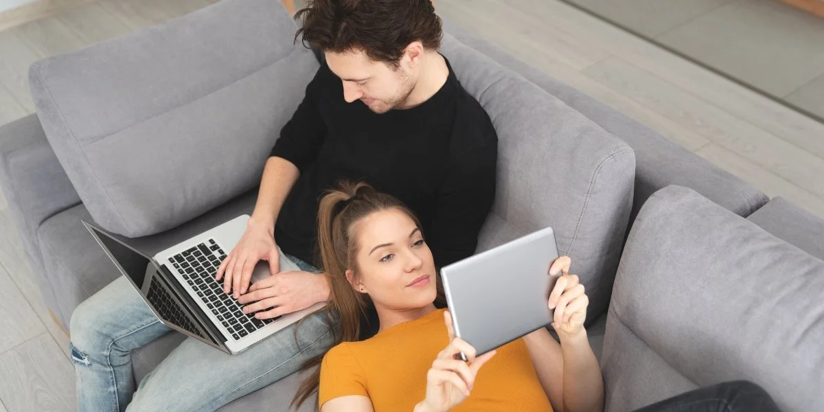 Two people are using a laptop and a tablet on a sofa
