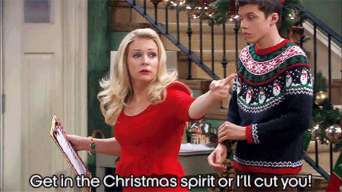 A woman in a Christmas setting encourages a man wearing a Christmas sweater to get into the Christmas spirit