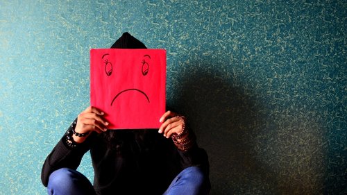 A person is holding a red sheet of paper with a sad face on it in front of her face.