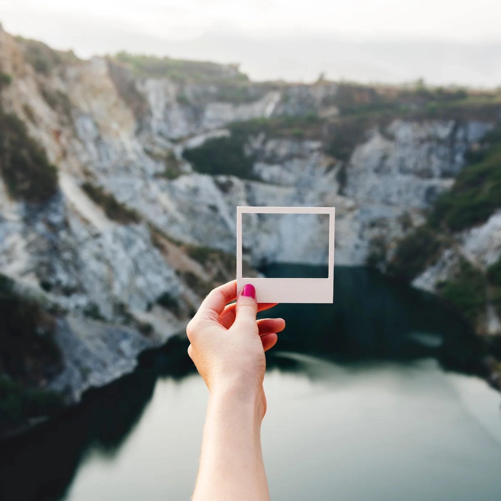 A hand holds an empty instant photo frame in front of a rocky landscape