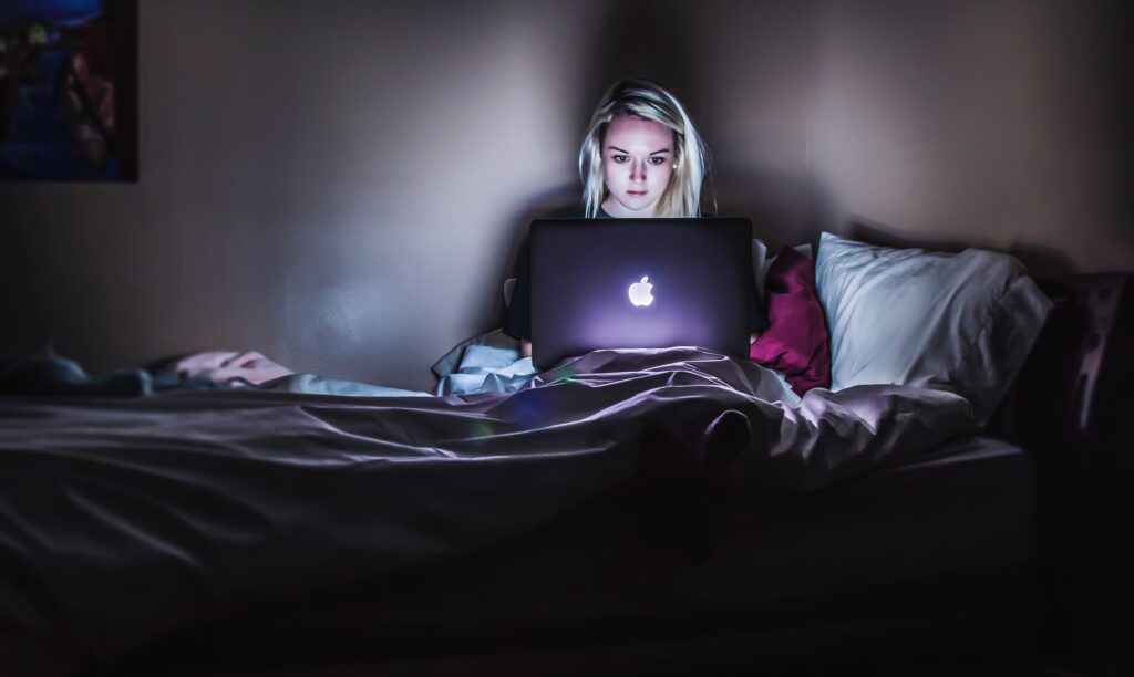 Une personne est assise dans son lit, la nuit, devant un ordinateur portable allumé.